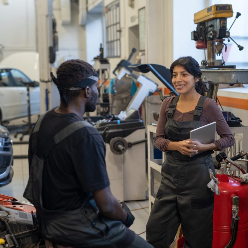 Vertical portrait of two smiling car mechanics chatting in garage shop, focus on young woman in workwear